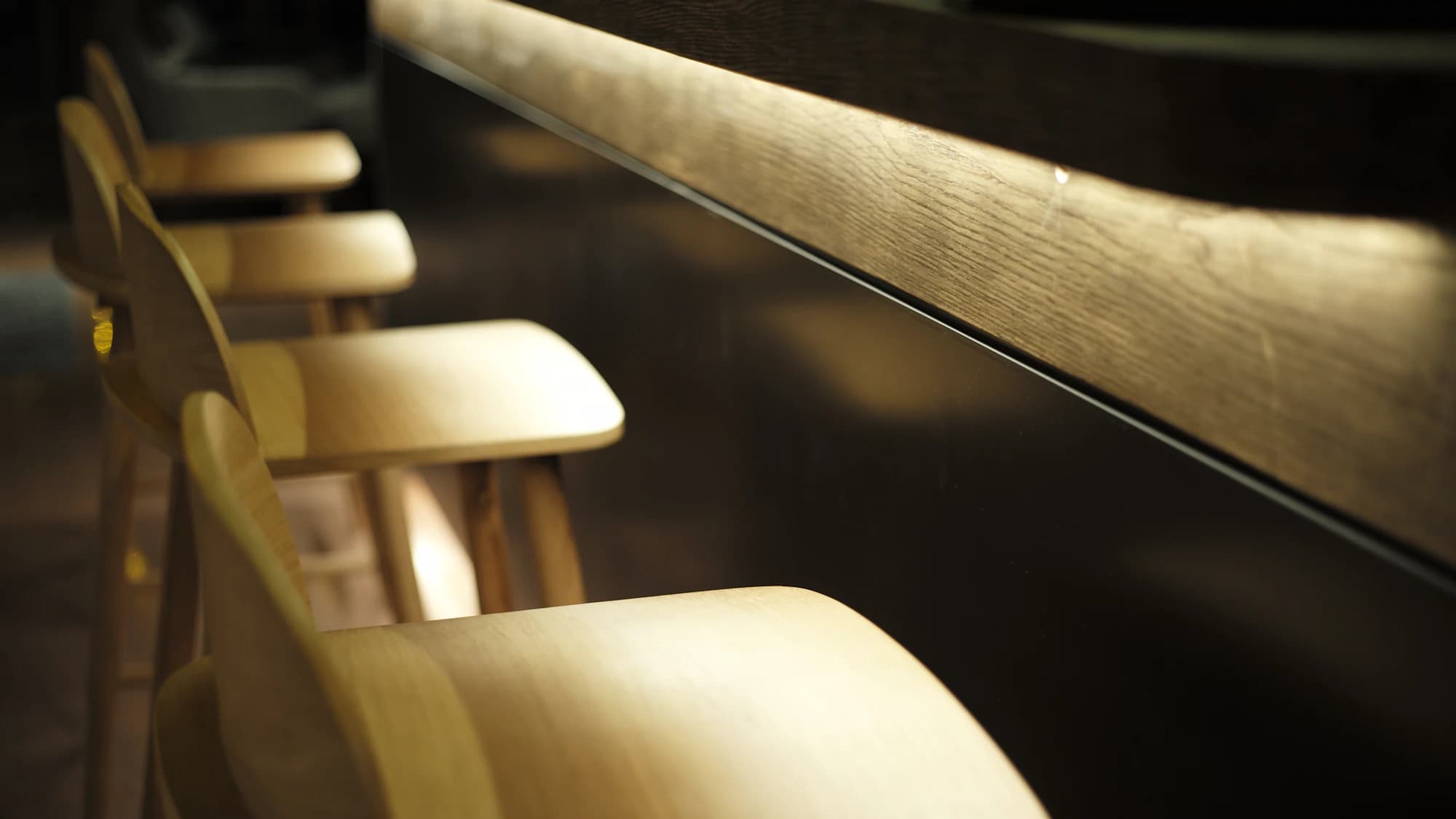 A row of pale wooden stools along the edge of a counter, low light raking across the timber.