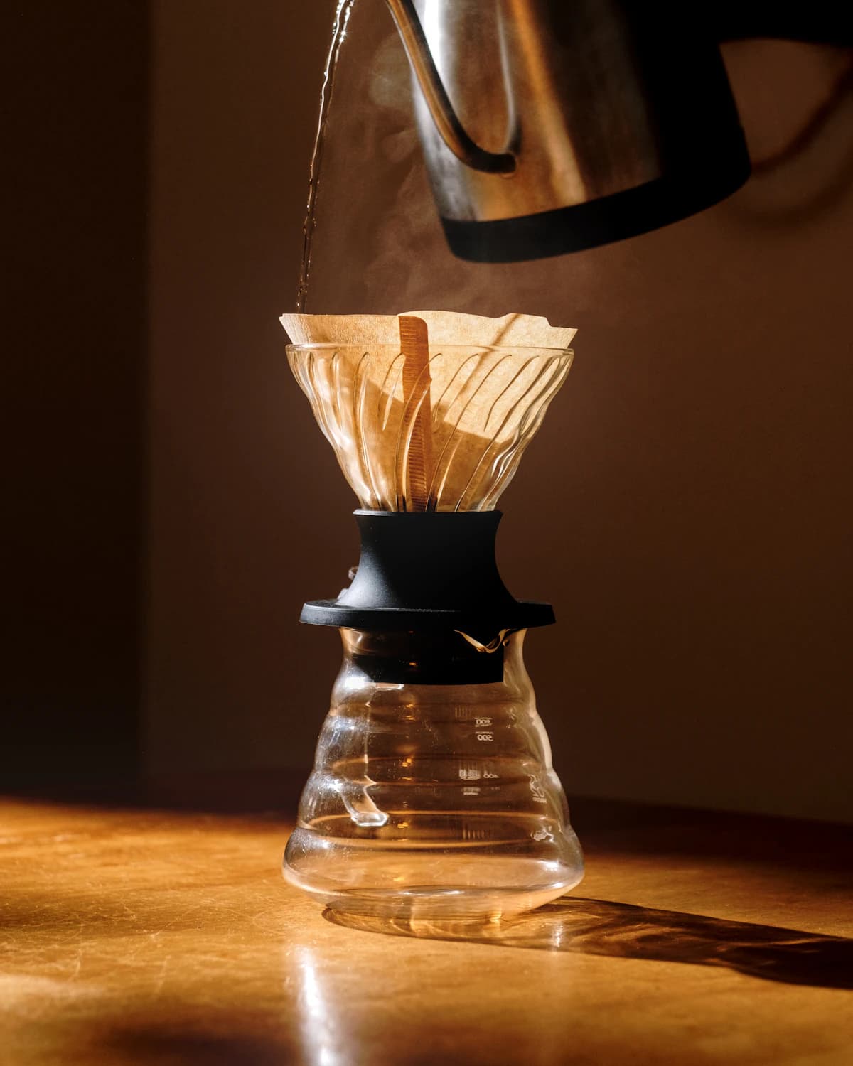 A gooseneck kettle pouring hot water through a paper filter into a glass carafe, lit from one side on a wooden counter.